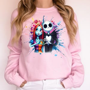 Op de afbeelding: Lichtroze sweatshirt met een kleurrijke afbeelding van Jack Skellington en Sally uit The Nightmare Before Christmas. De personages vormen een hart met hun handen, omringd door kleurrijke spetters.