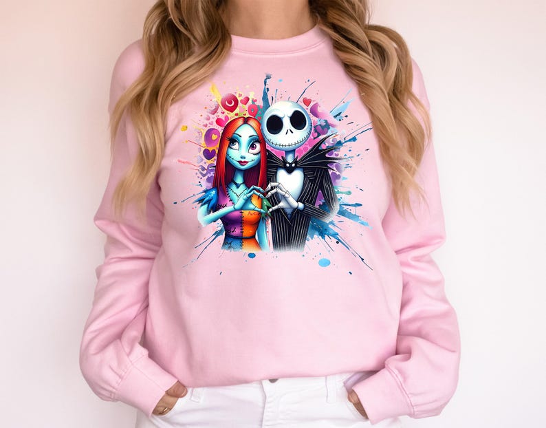 Op de afbeelding: Lichtroze sweatshirt met een kleurrijke afbeelding van Jack Skellington en Sally uit The Nightmare Before Christmas. De personages vormen een hart met hun handen, omringd door kleurrijke spetters.