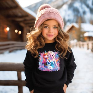 May include: A young girl wearing a black sweatshirt and a pink knit hat. The sweatshirt features a colorful graphic of animated characters. The background shows a snowy outdoor scene with wooden structures.