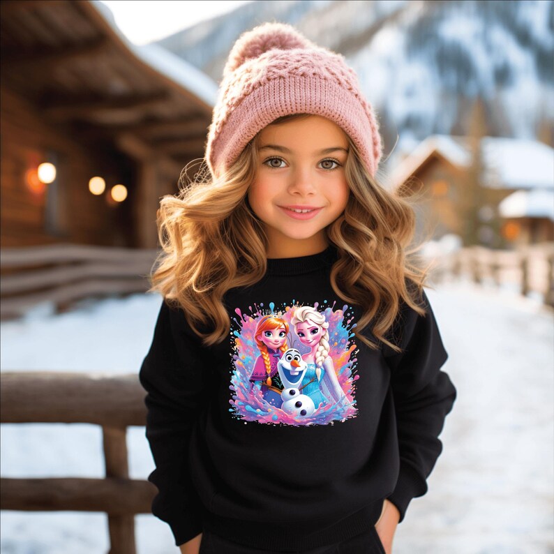 May include: A young girl wearing a black sweatshirt and a pink knit hat. The sweatshirt features a colorful graphic of animated characters. The background shows a snowy outdoor scene with wooden structures.