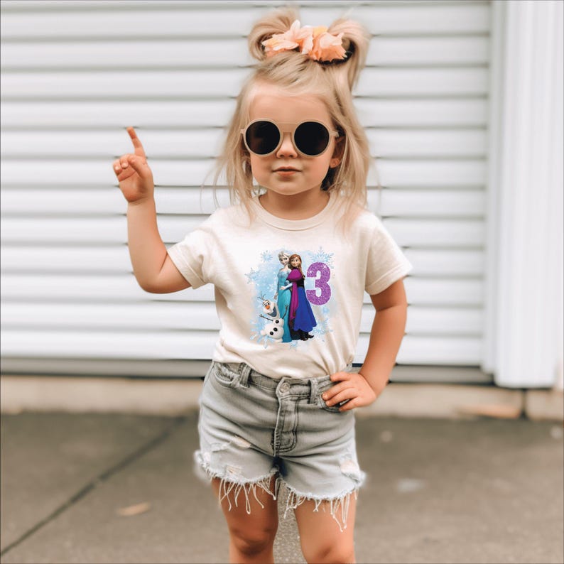 May include: A beige t-shirt featuring a graphic of characters from the movie Frozen, with the number 3 in purple. The shirt is worn by a young child wearing sunglasses and denim shorts.