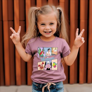 Op de afbeelding: Een jong meisje draagt een mauvekleurig T-shirt met een stripontwerp van "De Kleine Zeemeermin". Het shirt toont Ariel, Prins Eric, Koning Triton, Sebastiaan, Ursula en Botje. Het meisje lacht en maakt met beide handen een vredesteken.