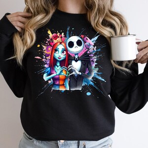 Op de afbeelding: Zwart sweatshirt met een kleurrijk aquarelontwerp van Jack Skellington en Sally uit The Nightmare Before Christmas. De personages vormen een hart met hun handen. Een witte mok wordt in de hand van de persoon gehouden.