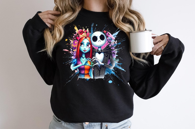 Op de afbeelding: Zwart sweatshirt met een kleurrijk aquarelontwerp van Jack Skellington en Sally uit The Nightmare Before Christmas. De personages vormen een hart met hun handen. Een witte mok wordt in de hand van de persoon gehouden.