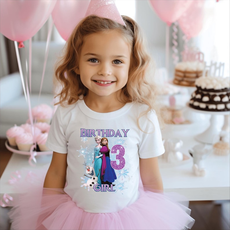 May include: A young girl wearing a white t-shirt with a "Birthday Girl 3" graphic featuring Elsa and Anna from Frozen. She is also wearing a pink tutu and a party hat, with pink balloons and cupcakes in the background.