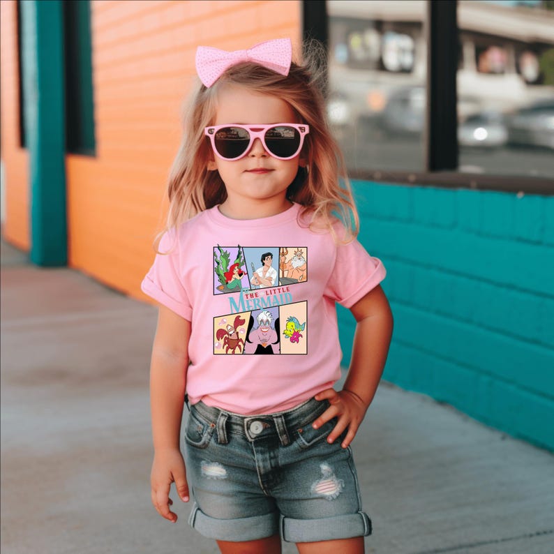 Op de afbeelding: Een roze T-shirt met een stripverhaalontwerp van "De Kleine Zeemeermin". Het shirt heeft een ronde hals en korte mouwen. Het ontwerp bevat Ariel, Prins Eric en andere personages. Het kind draagt een roze zonnebril en een roze strik.