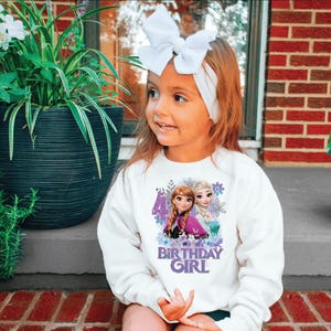 May include: A white sweatshirt featuring a graphic of two animated characters and the text "BIRTHDAY GIRL". The design includes the number 4 and floral accents. The girl is wearing a white bow headband.
