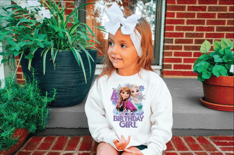 May include: A white sweatshirt featuring a graphic of two animated characters and the text "BIRTHDAY GIRL". The design includes the number 4 and floral accents. The girl is wearing a white bow headband.