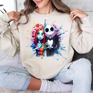 Op de afbeelding: Beige sweatshirt met een kleurrijke aquarel illustratie van Jack Skellington en Sally uit The Nightmare Before Christmas. Het kunstwerk bevat harten en spetters van blauw, roze en paars. De personages zijn afgebeeld in hun klassieke outfits.