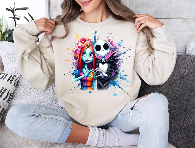 Op de afbeelding: Beige sweatshirt met een kleurrijke aquarel illustratie van Jack Skellington en Sally uit The Nightmare Before Christmas. Het kunstwerk bevat harten en spetters van blauw, roze en paars. De personages zijn afgebeeld in hun klassieke outfits.