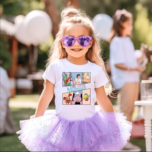 Op de afbeelding: Een jong meisje draagt een wit T-shirt met een stripontwerp van "De Kleine Zeemeermin" en een paarse tutu. Het T-shirt-ontwerp bevat Ariel, Prins Eric, Koning Triton, Sebastiaan, Ursula en Botje. Ze draagt ook een paarse zonnebril.
