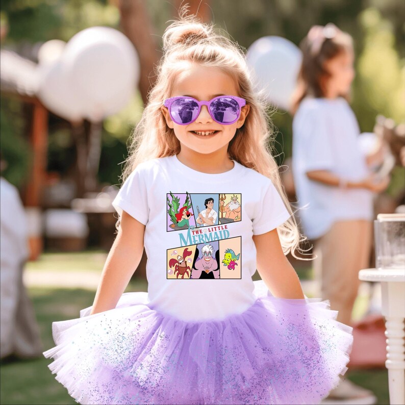 Op de afbeelding: Een jong meisje draagt een wit T-shirt met een stripontwerp van "De Kleine Zeemeermin" en een paarse tutu. Het T-shirt-ontwerp bevat Ariel, Prins Eric, Koning Triton, Sebastiaan, Ursula en Botje. Ze draagt ook een paarse zonnebril.