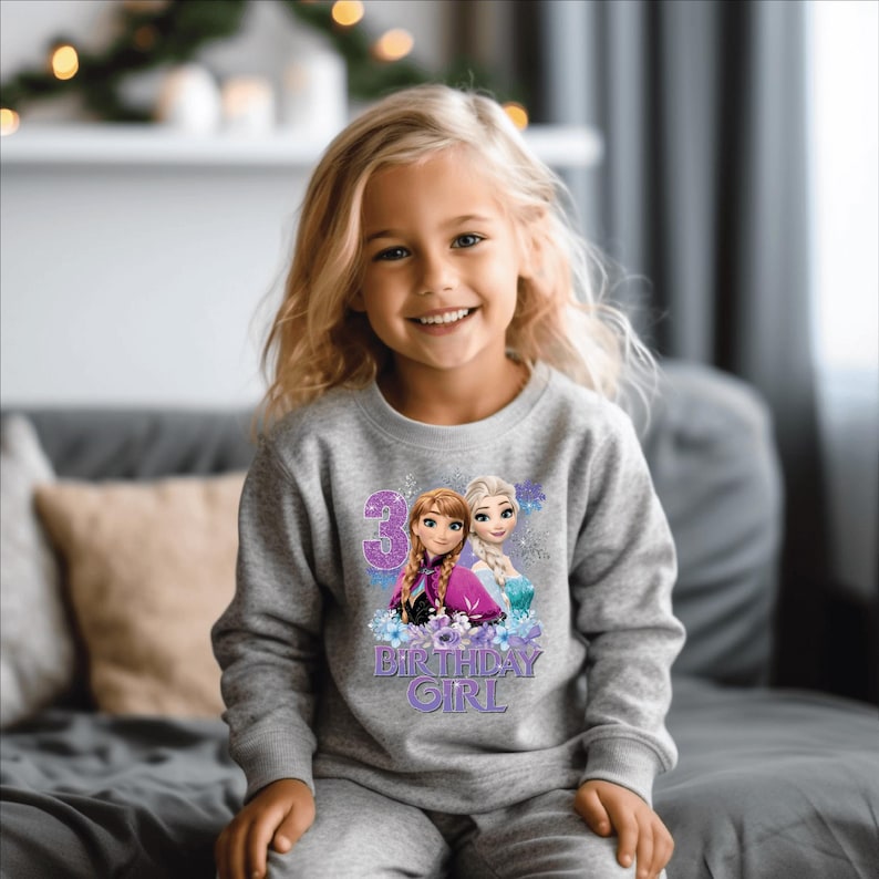 May include: A gray sweatshirt and matching pants set featuring a graphic of Anna and Elsa from Frozen, with the number 3 and the words "Birthday Girl". The sweatshirt is worn by a young girl with blonde hair.