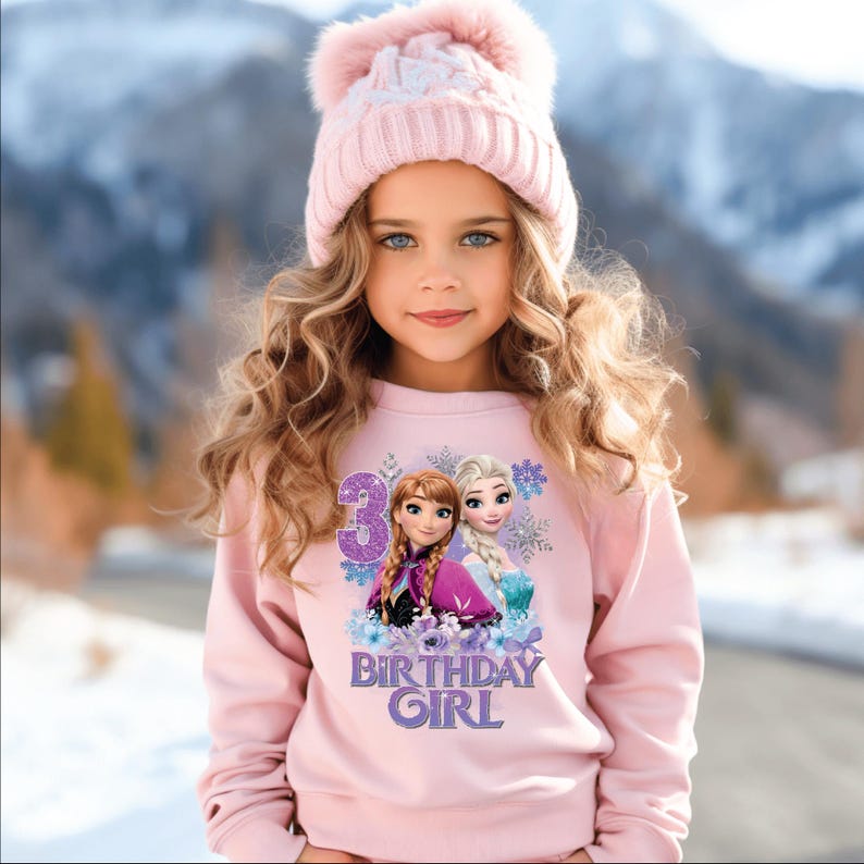 May include: A light pink sweatshirt featuring a graphic of Anna and Elsa from Frozen, with the number 3 and the text "BIRTHDAY GIRL". The sweatshirt is paired with a matching pink knit hat with a pom-pom.