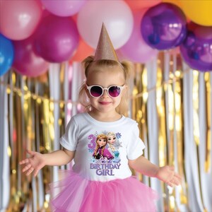 May include: A young girl wearing a white t-shirt with a graphic of two cartoon characters and the text "3 BIRTHDAY GIRL". She is wearing a pink tutu, sunglasses, and a gold party hat. Balloons and streamers are in the background.