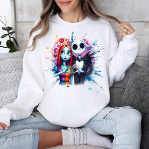 Op de afbeelding: Witte sweatshirt met een kleurrijk aquarelontwerp van Jack Skellington en Sally uit The Nightmare Before Christmas. De afbeelding toont de personages die elkaars handen vasthouden, omringd door harten en verfspatten. Een casual top met lange mouwen.