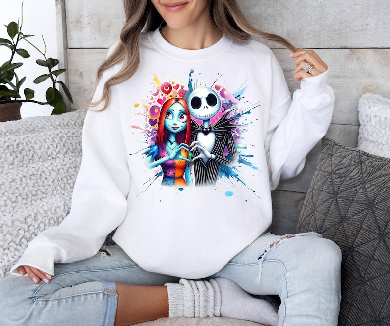 Op de afbeelding: Witte sweatshirt met een kleurrijk aquarelontwerp van Jack Skellington en Sally uit The Nightmare Before Christmas. De afbeelding toont de personages die elkaars handen vasthouden, omringd door harten en verfspatten. Een casual top met lange mouwen.
