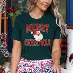 Op de afbeelding: Donkergroen t-shirt met de tekst "MERRY CHRISTMAS" in rode blokletters. Een vintage illustratie van de kerstman staat eronder. Het shirt wordt gedragen met een rood-witte pyjamabroek met patroon.