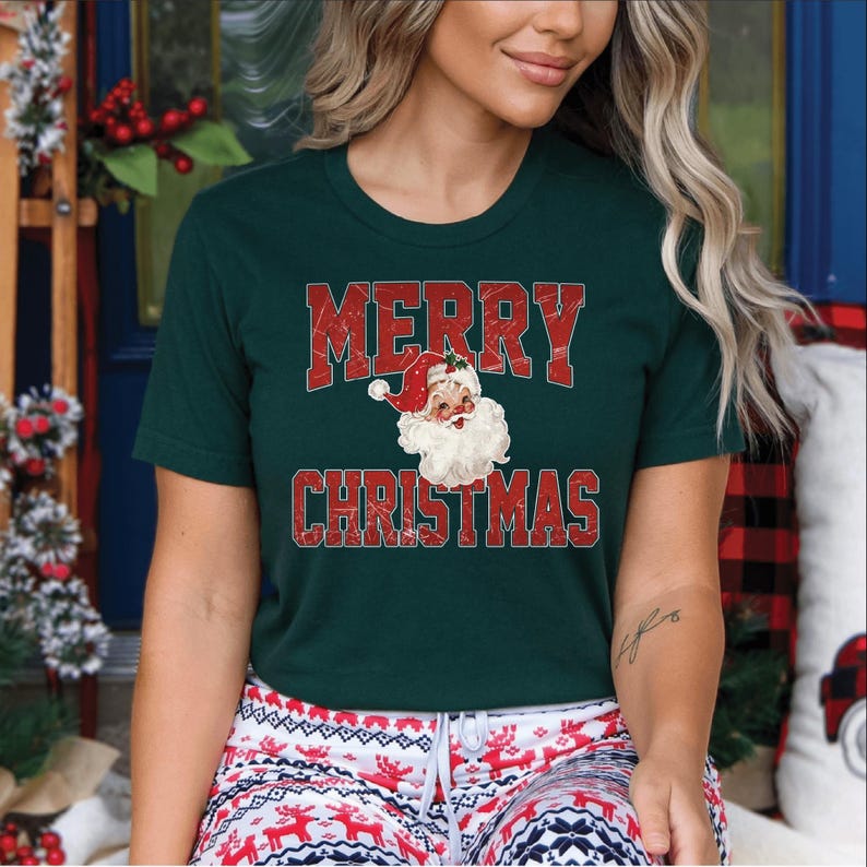 Op de afbeelding: Donkergroen t-shirt met de tekst "MERRY CHRISTMAS" in rode blokletters. Een vintage illustratie van de kerstman staat eronder. Het shirt wordt gedragen met een rood-witte pyjamabroek met patroon.