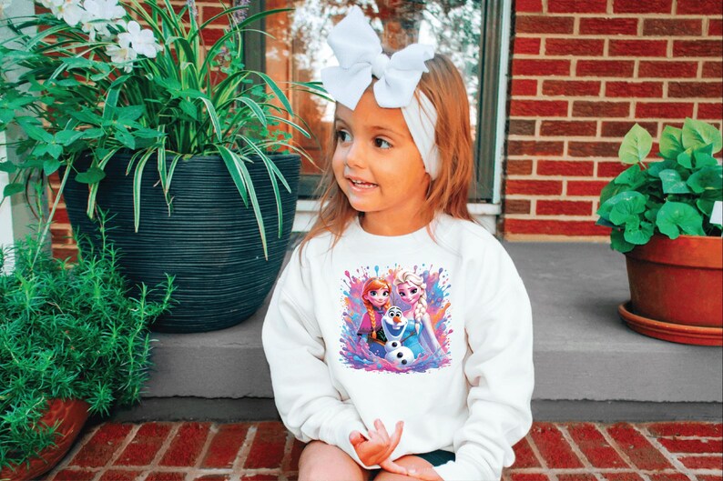 May include: A young girl wearing a white sweatshirt featuring a colorful graphic of characters from the movie "Frozen". She has a white bow headband and is sitting on a brick surface. Potted plants are in the background.