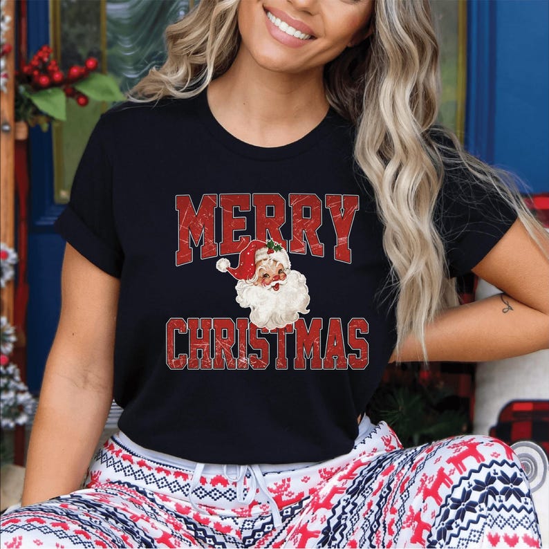 Op de afbeelding: Zwart T-shirt met de woorden "MERRY CHRISTMAS" in rode blokletters. Het shirt heeft een Santa Claus-afbeelding in het midden. De persoon draagt een rood-witte pyjamabroek met patroon.