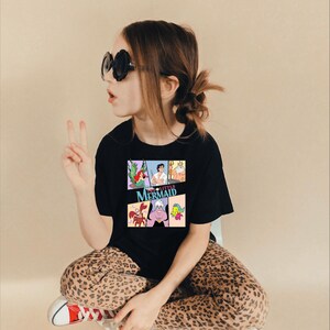 Op de afbeelding: Zwart T-shirt met een kleurrijke afbeelding van personages uit de film "De Kleine Zeemeermin". Het ontwerp bevat Ariel, Prins Eric, Koning Triton, Ursula, Sebastiaan en Botje. Het T-shirt wordt gedragen door een persoon met een zonnebril en een luipaardbroek.