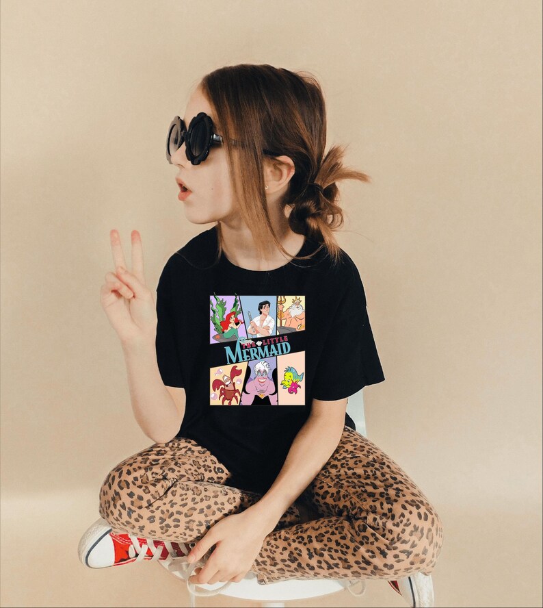 Op de afbeelding: Zwart T-shirt met een kleurrijke afbeelding van personages uit de film "De Kleine Zeemeermin". Het ontwerp bevat Ariel, Prins Eric, Koning Triton, Ursula, Sebastiaan en Botje. Het T-shirt wordt gedragen door een persoon met een zonnebril en een luipaardbroek.