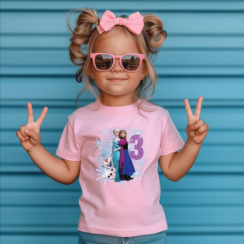 May include: A pink t-shirt featuring a graphic of characters from the movie "Frozen" and the number "3". The shirt is worn by a young child wearing pink sunglasses and a pink bow. The background is a blue wall.