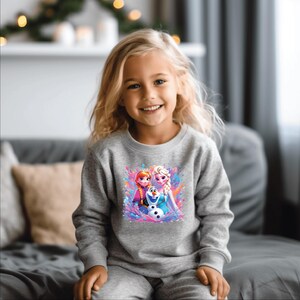 May include: A gray sweatshirt featuring a colorful graphic of two princesses and a snowman. The long-sleeved top is worn by a smiling child. The graphic has a watercolor effect with pink, blue, and purple hues.