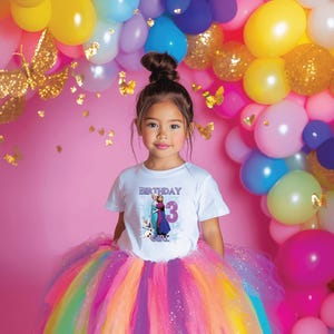 May include: A young girl wearing a white t-shirt with a graphic of two characters and the number 3, with the word "BIRTHDAY" above and "GIRL" below. She is wearing a colorful rainbow tutu skirt. The background includes balloons and butterflies.