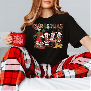 Puede incluir: Camiseta negra con un dise&ntilde;o navide&ntilde;o colorido con personajes de Disney, incluyendo Mickey y Minnie Mouse. La palabra "CHRISTMAS" est&aacute; escrita en la parte superior. Se sostiene una taza roja con el texto "Have yourself a Merry Little Christmas".