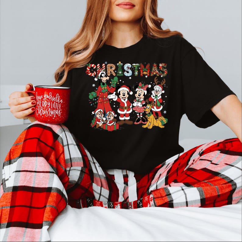 Puede incluir: Camiseta negra con un dise&ntilde;o navide&ntilde;o colorido con personajes de Disney, incluyendo Mickey y Minnie Mouse. La palabra "CHRISTMAS" est&aacute; escrita en la parte superior. Se sostiene una taza roja con el texto "Have yourself a Merry Little Christmas".