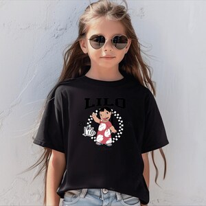 Puede incluir: Camiseta negra con la palabra "LILO" encima de una imagen de dibujos animados de una ni&ntilde;a con un vestido rojo con lunares blancos. La imagen est&aacute; rodeada por un c&iacute;rculo a cuadros blanco y negro. La camiseta tambi&eacute;n tiene el texto "2002" y un signo de mano.