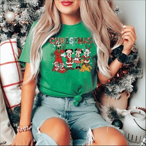Puede incluir: Camiseta navide&ntilde;a verde con un gr&aacute;fico festivo de personajes de Disney, incluyendo Mickey y Minnie Mouse, Goofy y Chip y Dale, con atuendos navide&ntilde;os. La palabra "CHRISTMAS" est&aacute; impresa en la parte superior en una fuente colorida y juguetona. La camiseta est&aacute; anudada en la cintura.