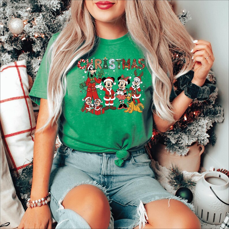 Puede incluir: Camiseta navide&ntilde;a verde con un gr&aacute;fico festivo de personajes de Disney, incluyendo Mickey y Minnie Mouse, Goofy y Chip y Dale, con atuendos navide&ntilde;os. La palabra "CHRISTMAS" est&aacute; impresa en la parte superior en una fuente colorida y juguetona. La camiseta est&aacute; anudada en la cintura.