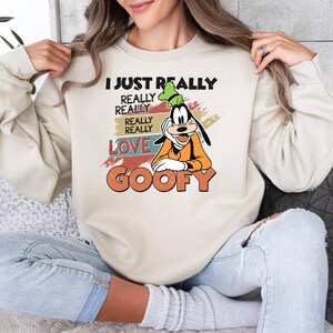 May include: Cream-colored sweatshirt featuring a graphic of Goofy with the text "I JUST REALLY REALLY REALLY LOVE GOOFY" in black, orange, and red. The design has a vintage aesthetic. The sweatshirt is a casual, comfortable style.