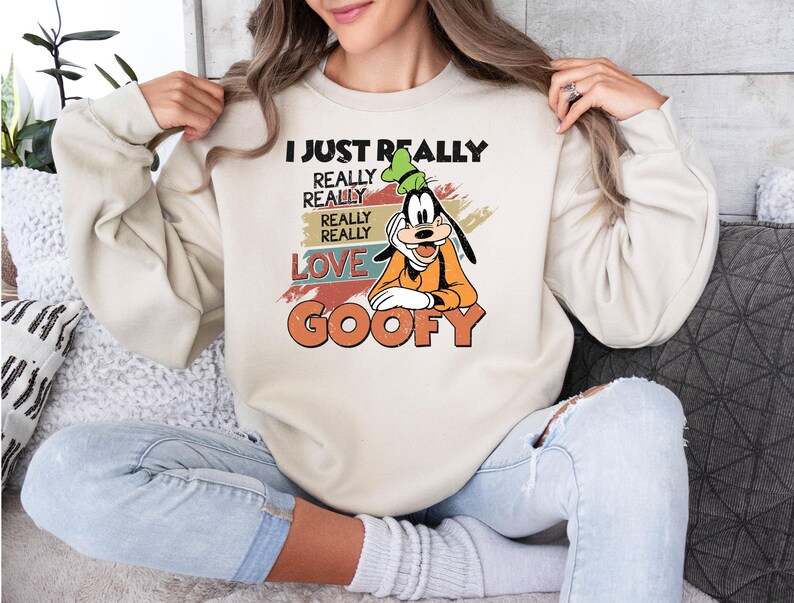 May include: Cream-colored sweatshirt featuring a graphic of Goofy with the text "I JUST REALLY REALLY REALLY LOVE GOOFY" in black, orange, and red. The design has a vintage aesthetic. The sweatshirt is a casual, comfortable style.