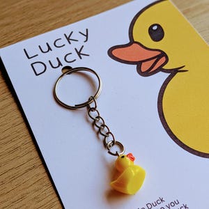 May include: A yellow rubber duck keychain with a red beak and a silver chain attached to a silver key ring. The keychain is on a white card with the text "Lucky Duck" and a cartoon duck illustration. The card reads "A little Duck from me to you to bring you luck in all you do."