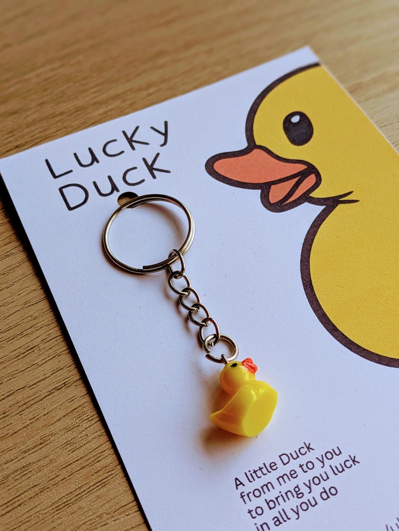 May include: A yellow rubber duck keychain with a red beak and a silver chain attached to a silver key ring. The keychain is on a white card with the text "Lucky Duck" and a cartoon duck illustration. The card reads "A little Duck from me to you to bring you luck in all you do."