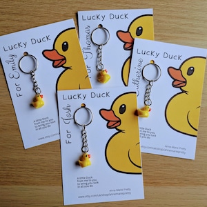 May include: Four white cards with yellow rubber duck keychains attached. Each card has the text "Lucky Duck" and a name, "For Emily", "For Thomas", "For Josh", and "Catherine". The cards have a message about the duck bringing good luck.