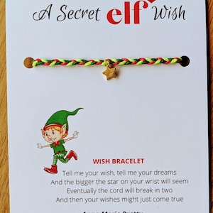 May include: A Secret Elf Wish bracelet on a white card. The bracelet features a braided cord in green, pink, and yellow, with a small gold star charm. The card includes an elf illustration and text about wishes.