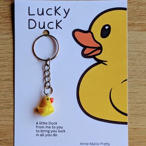 May include: A yellow rubber duck keychain with a silver chain and a ring. The duck has a red beak and orange feet. The keychain comes with a card that says "Lucky Duck" and "A little Duck from me to you to bring you luck in all you do." The card also includes the name "Anne-Marie Pretty" and the website "www.etsy.com/uk/shop/annemariepretty".