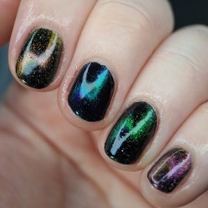May include: Close-up of four fingernails painted with black nail polish with green, blue, and purple iridescent glitter. The nail polish has a magnetic effect, creating a swirling pattern.