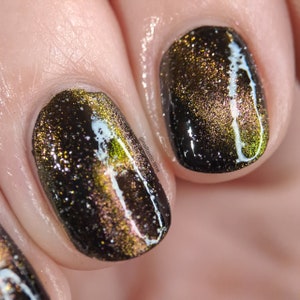 May include: Close-up of two fingernails painted with a dark, glittery, iridescent black nail polish with white lines. The polish has a green and gold shimmer.