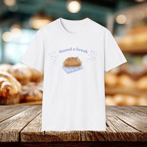 May include: White t-shirt with the text "Knead a break" above a cartoon loaf of bread on a blue checkered cloth. Small blue hearts are scattered around the text and bread illustration.