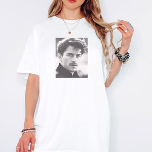 May include: White t-shirt featuring a black and white portrait of a man. The portrait is centered on the front of the shirt. The shirt has short sleeves and a crew neck.