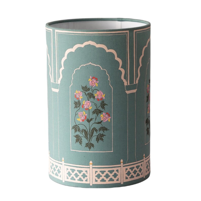 May include: A teal blue cylindrical lampshade with a white and pink floral design. The design features a floral motif with pink flowers and green leaves. The lampshade has a white border with a decorative pattern.