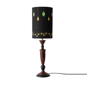 May include: A black table lamp with a dark wood base and a black fabric lampshade with a gold string of lights and hanging lanterns design.
