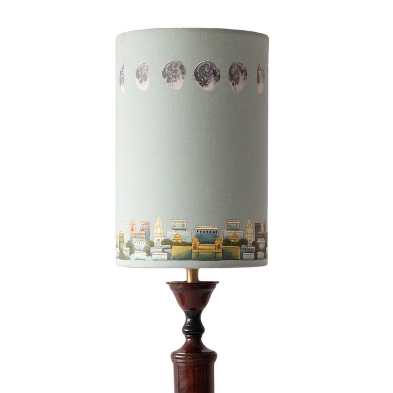 May include: A light blue lampshade with a cityscape and moon phases design. The lampshade is on a dark brown wooden lamp base.