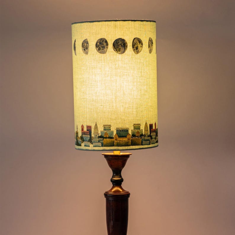 May include: A beige lampshade with a cityscape and moon phases design. The lampshade is on a brown wooden lamp base.